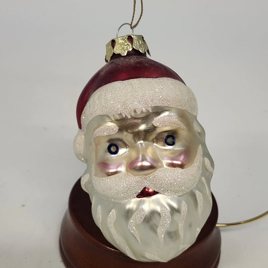 Blown glass Santa face ornament with red hat, glittered white beard, and painted facial details.