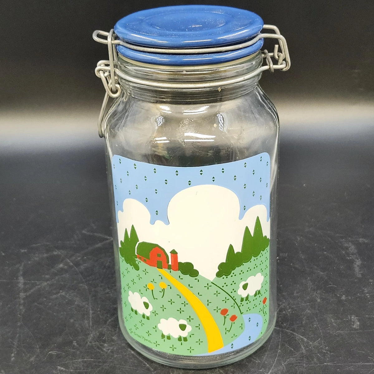 Vintage 1982 Anchor Hocking glass canister with colorful Nina farm scene, blue lid, and metal clamp closure.