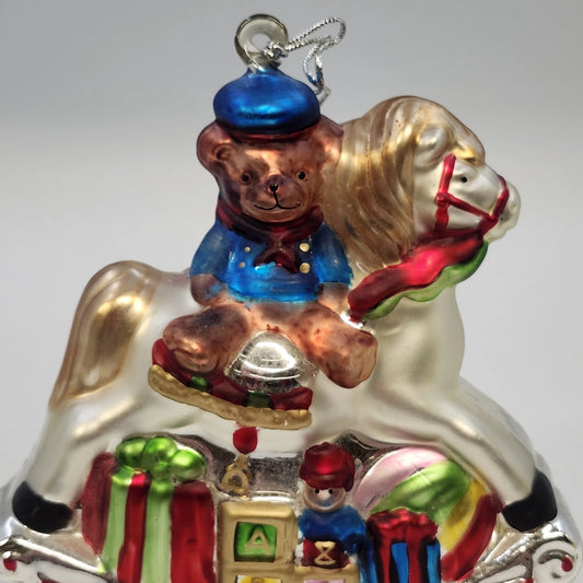 Vintage glass Christmas ornament featuring a teddy bear riding a white rocking horse with colorful toys and presents