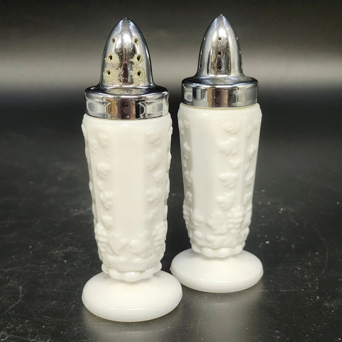 Pair of white milk glass salt and pepper shakers with raised grape and vine designs and silver-tone metal tops.